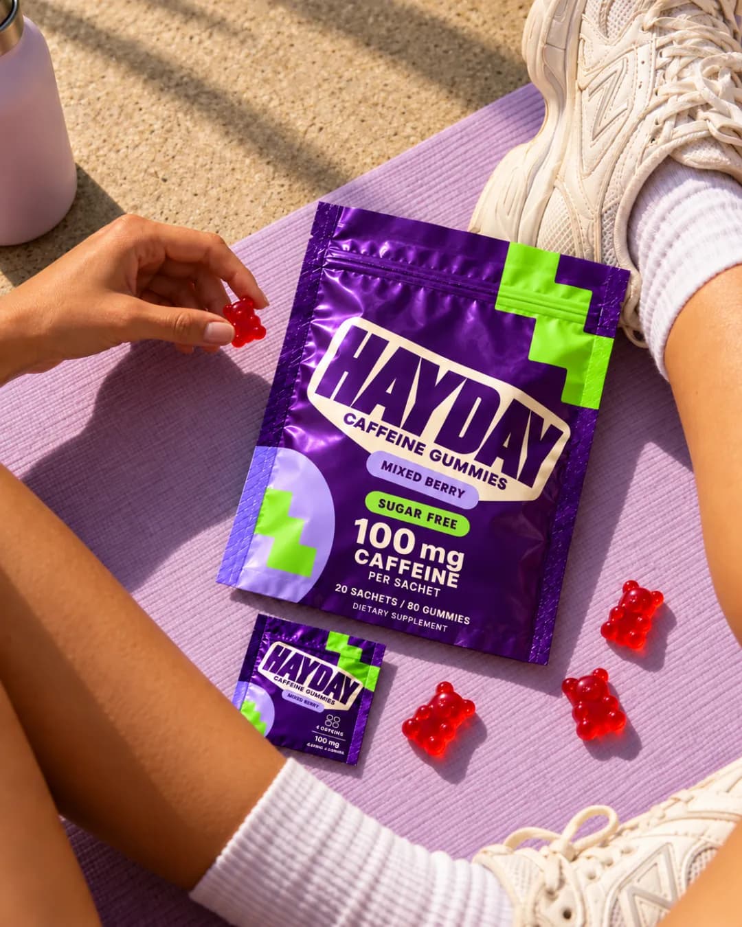 HAYDAY Mixed Berry pouch, a sachet and red gummies on a lilac yoga mat, a hand picking up a gummy